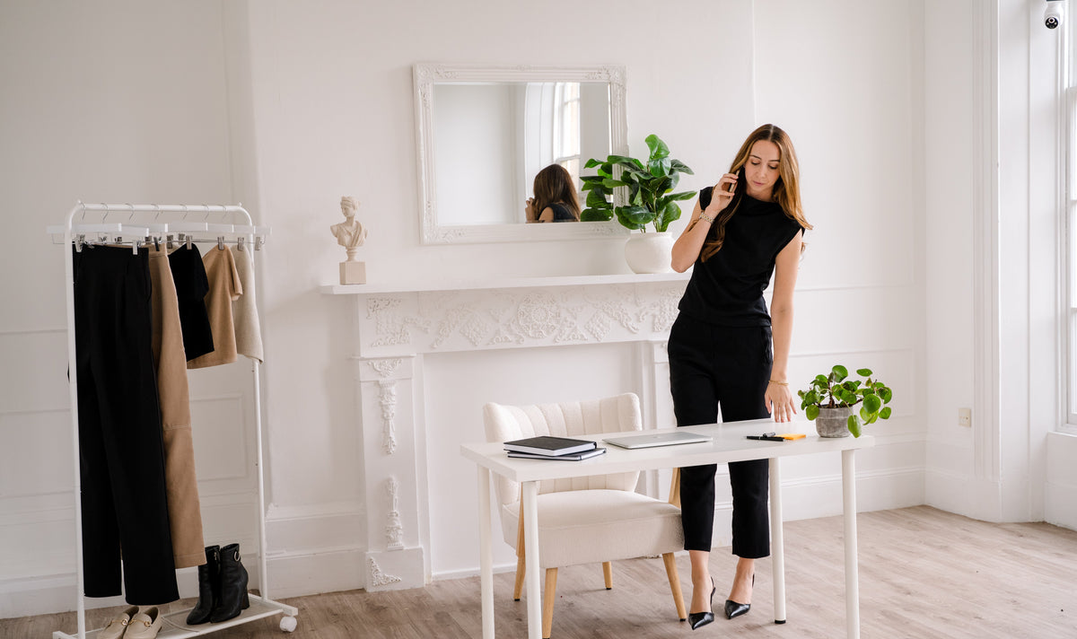 A woman in a black outfit stands in a bright, modern room next to a white desk, holding a phone to her ear. To her left is a clothing rack with black and beige pants, and the room features a white fireplace mantel, mirror, and light wood floors.