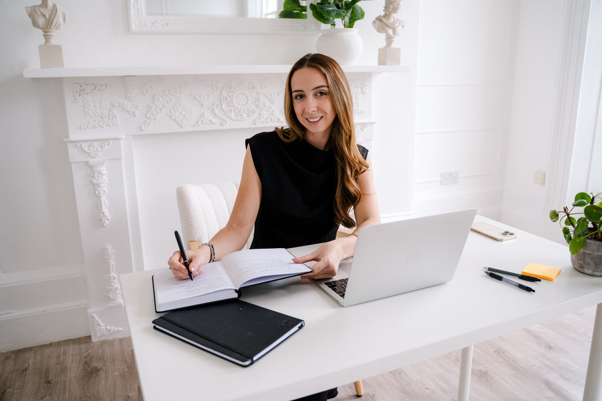 A woman with long brown hair sits at a white desk in a bright, modern room, smiling while writing in a notebook with a pen. A silver laptop, a black notebook, and other office items are on the desk in front of her.