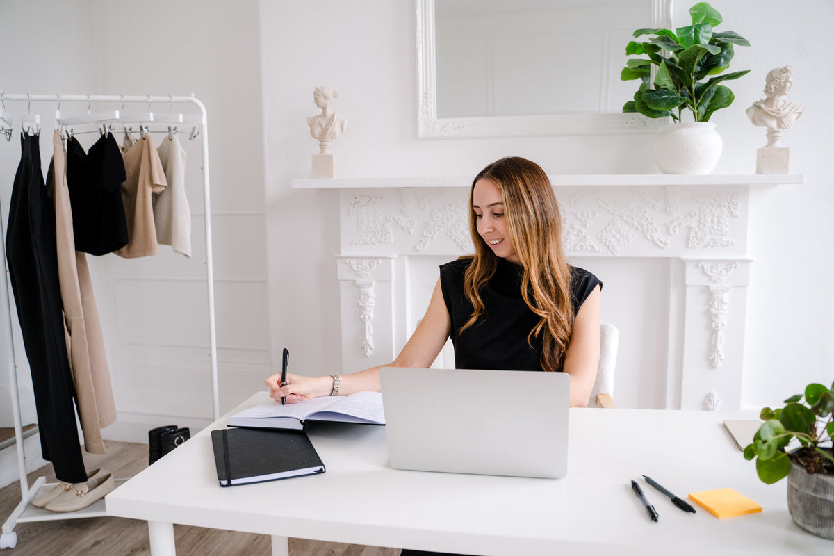A woman with long light brown hair sits at a white desk in a bright, modern room, writing in a notebook with a pen. She is wearing a black sleeveless top.