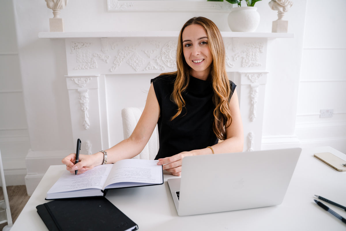 A woman with long light brown hair sits at a white desk in front of a white fireplace, wearing a black sleeveless top. She is smiling and holding a pen over an open notebook, with a silver laptop in front of her.