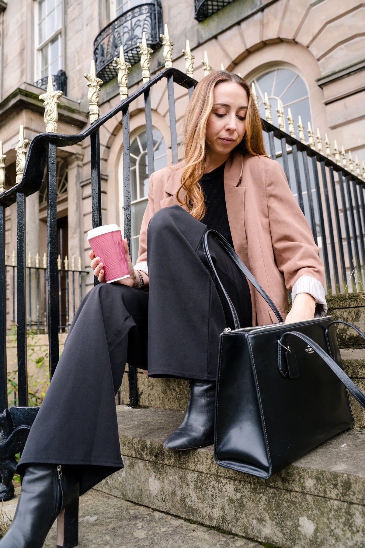 A woman sits on stone steps in front of a historic building, holding a pink coffee cup and wearing a dusty pink blazer, black wide-leg trousers, and black boots. A large black leather handbag rests on the step beside her.